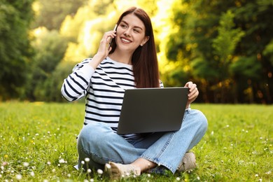 Photo of Smiling freelancer with laptop talking by smartphone on green grass outdoors. Remote job