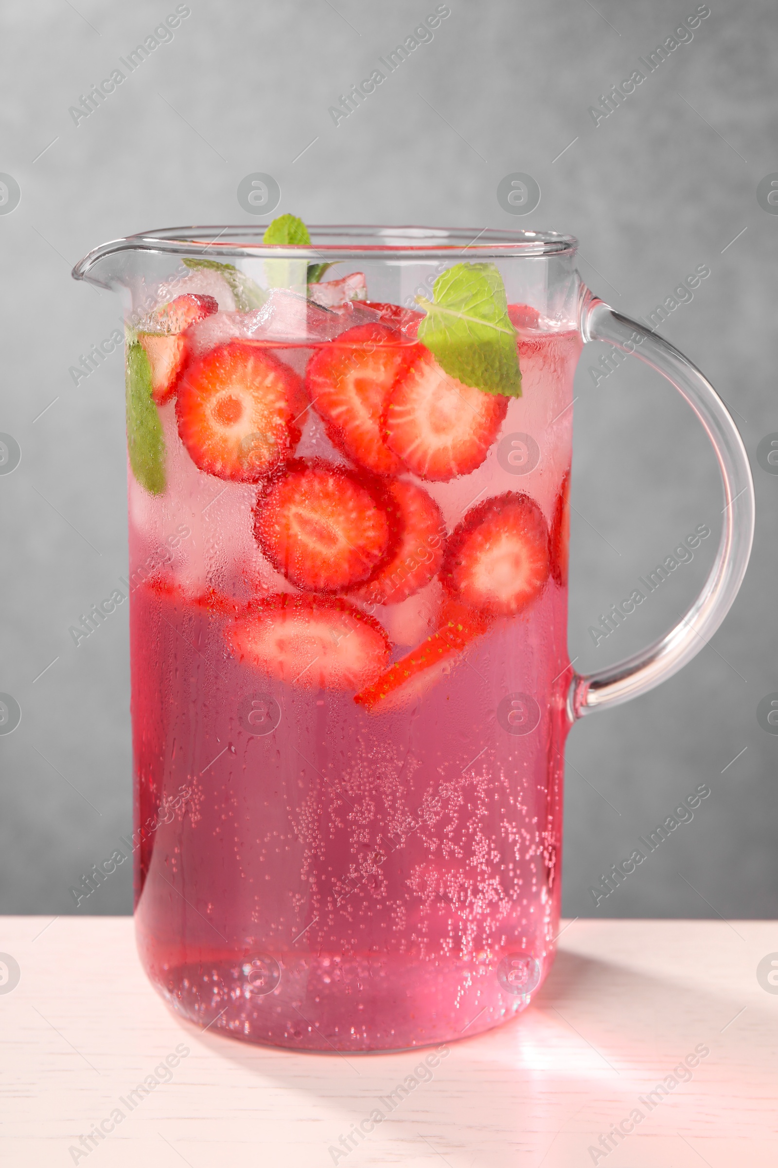 Photo of Freshly made strawberry lemonade with mint in jug and glass on white wooden table against grey background