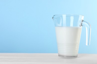 Photo of Jug of fresh milk on white wooden table against light blue background, space for text
