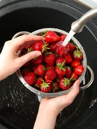 Photo of Woman washing fresh strawberries under tap water in metal colander above sink, top view