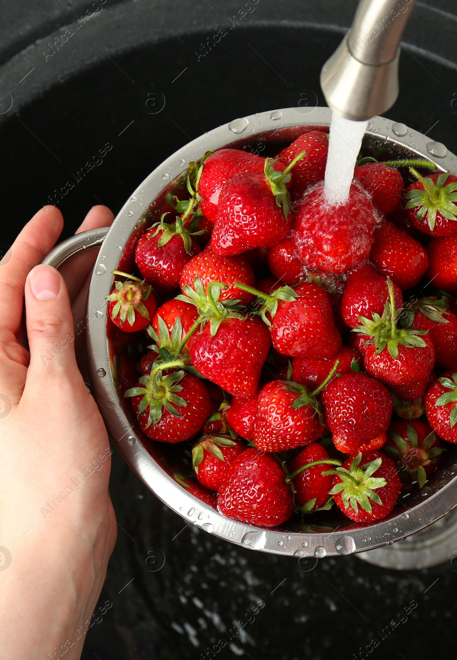 Photo of Woman washing fresh strawberries under tap water in metal colander above sink, top view