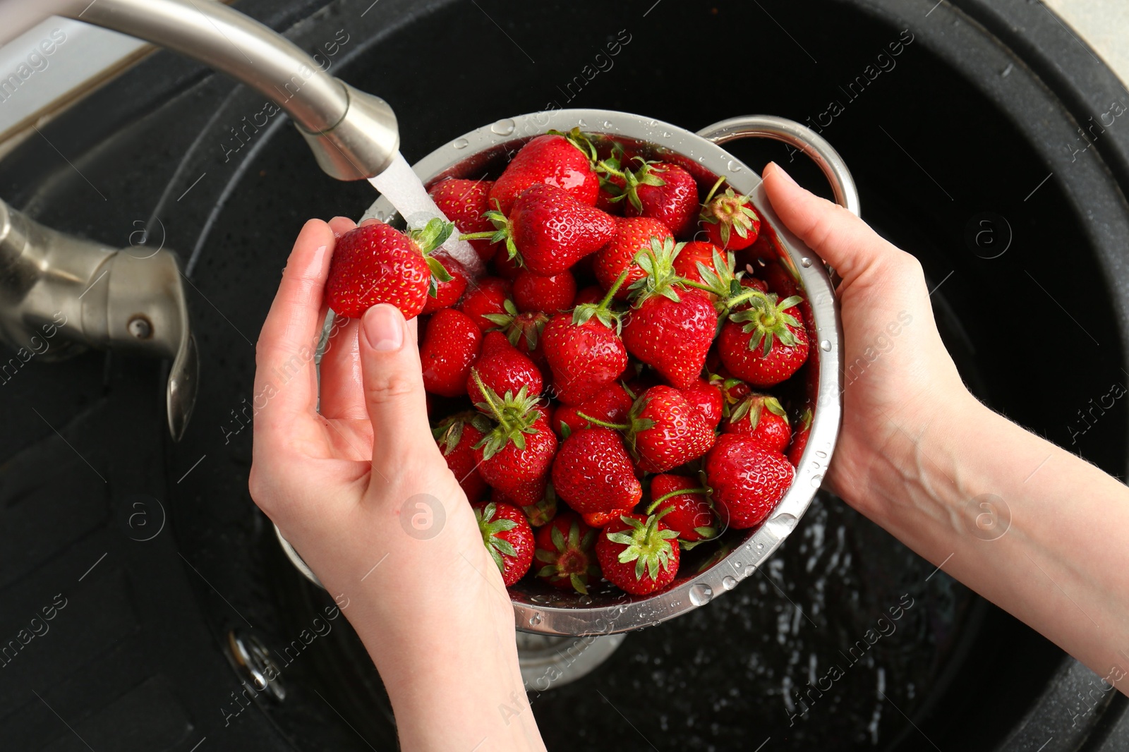 Photo of Woman washing fresh strawberries under tap water in metal colander above sink, top view