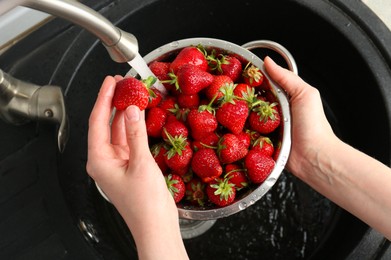 Photo of Woman washing fresh strawberries under tap water in metal colander above sink, top view