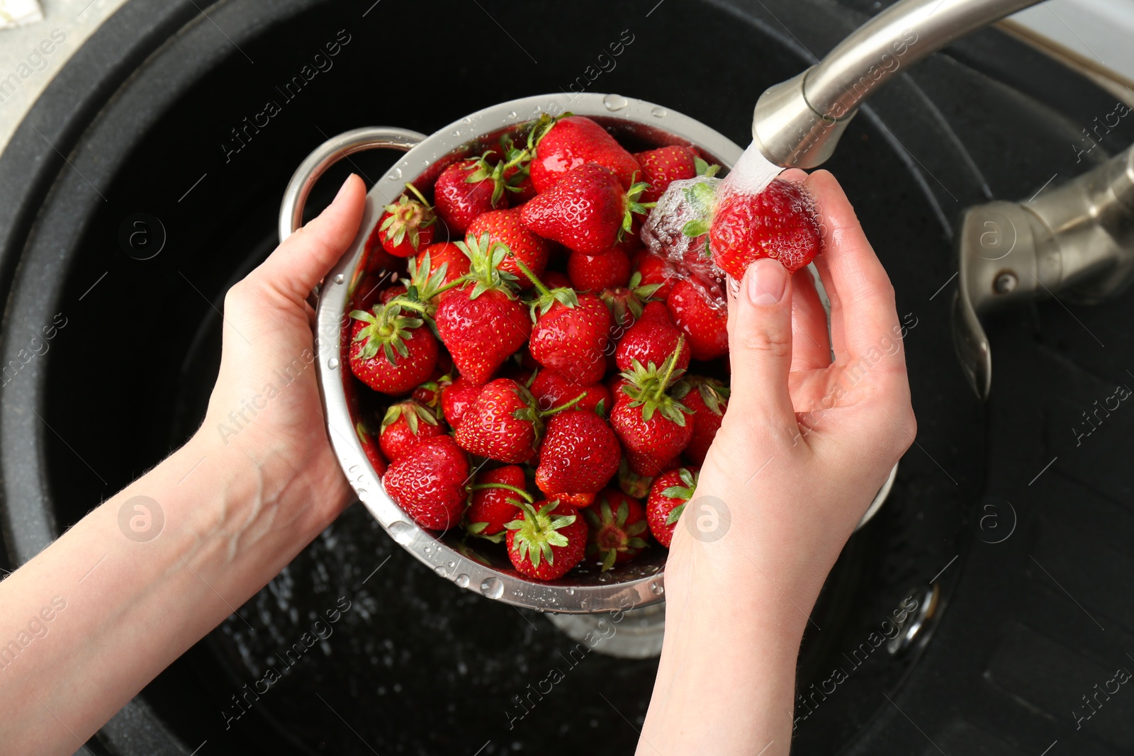 Photo of Woman washing fresh strawberries under tap water in metal colander above sink, top view