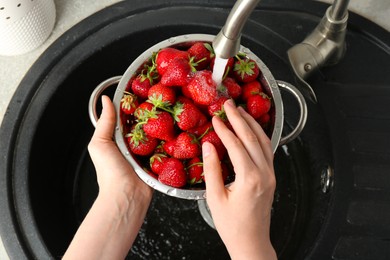 Photo of Woman washing fresh strawberries under tap water in metal colander above sink, top view