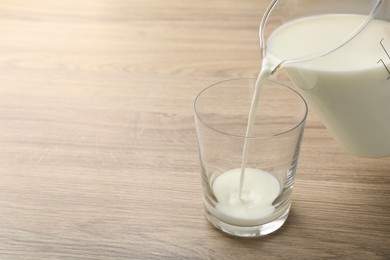Photo of Pouring fresh milk from jug into glass at wooden table, closeup. Space for text