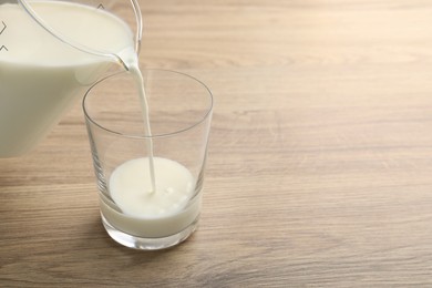 Photo of Pouring fresh milk from jug into glass at wooden table, closeup. Space for text