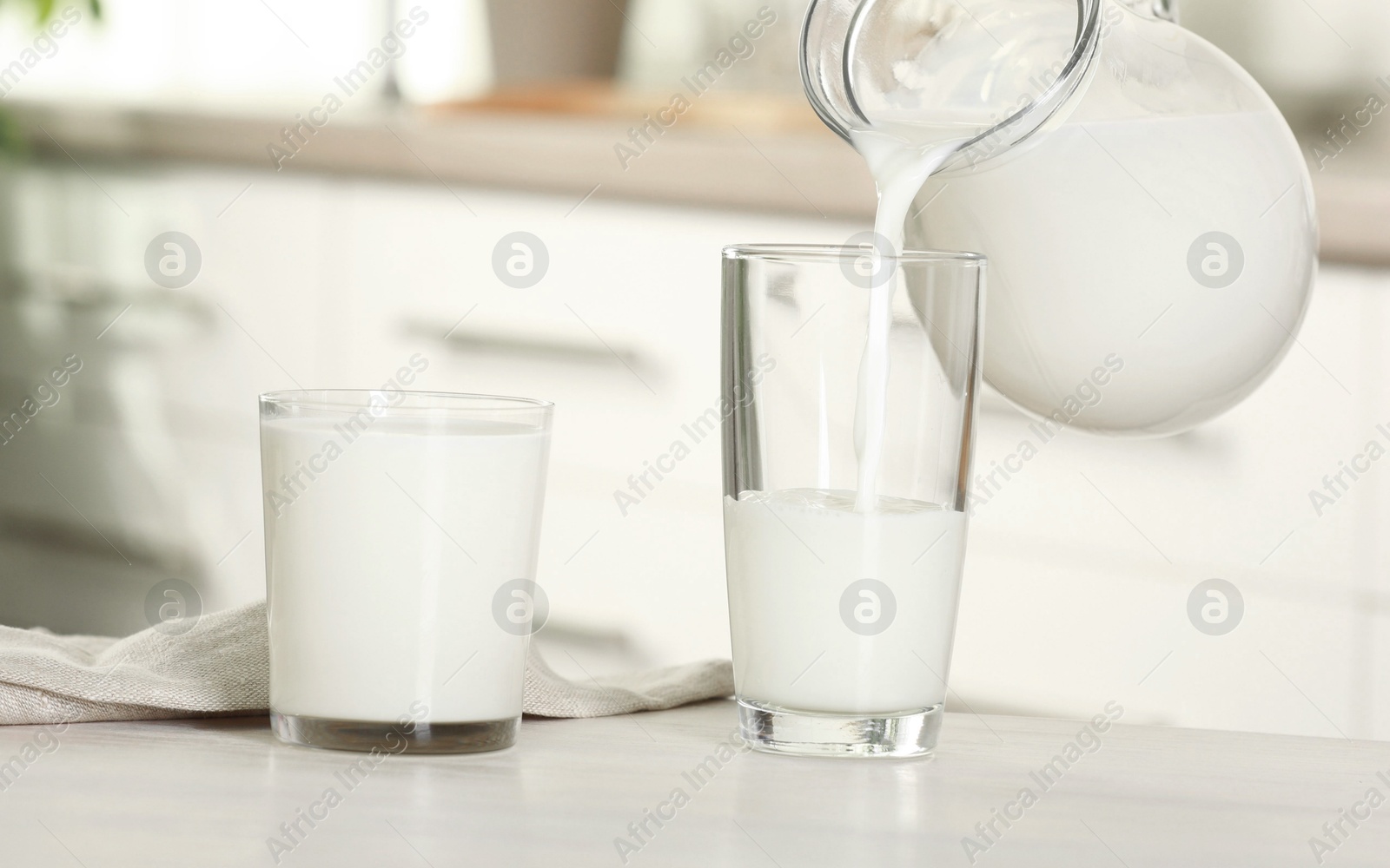 Photo of Pouring fresh milk from jug into glass at light table in kitchen, closeup
