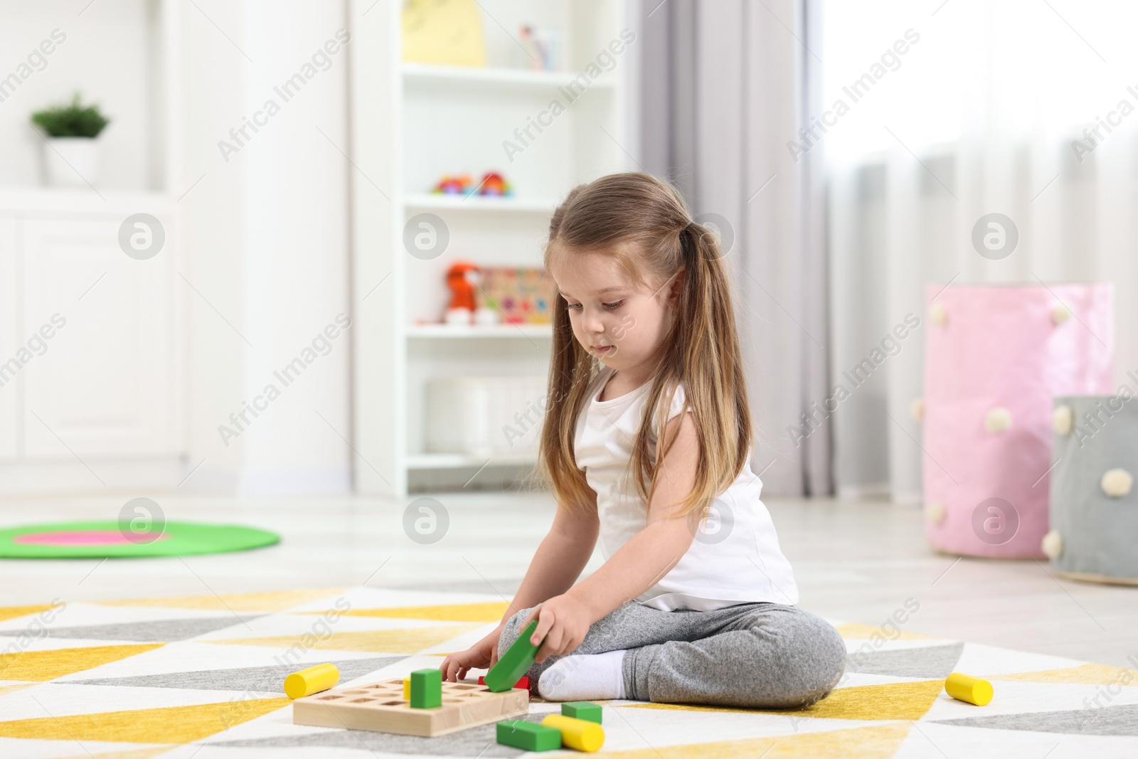 Photo of Cute little girl playing with set of wooden geometric figures on floor in kindergarten. Space for text