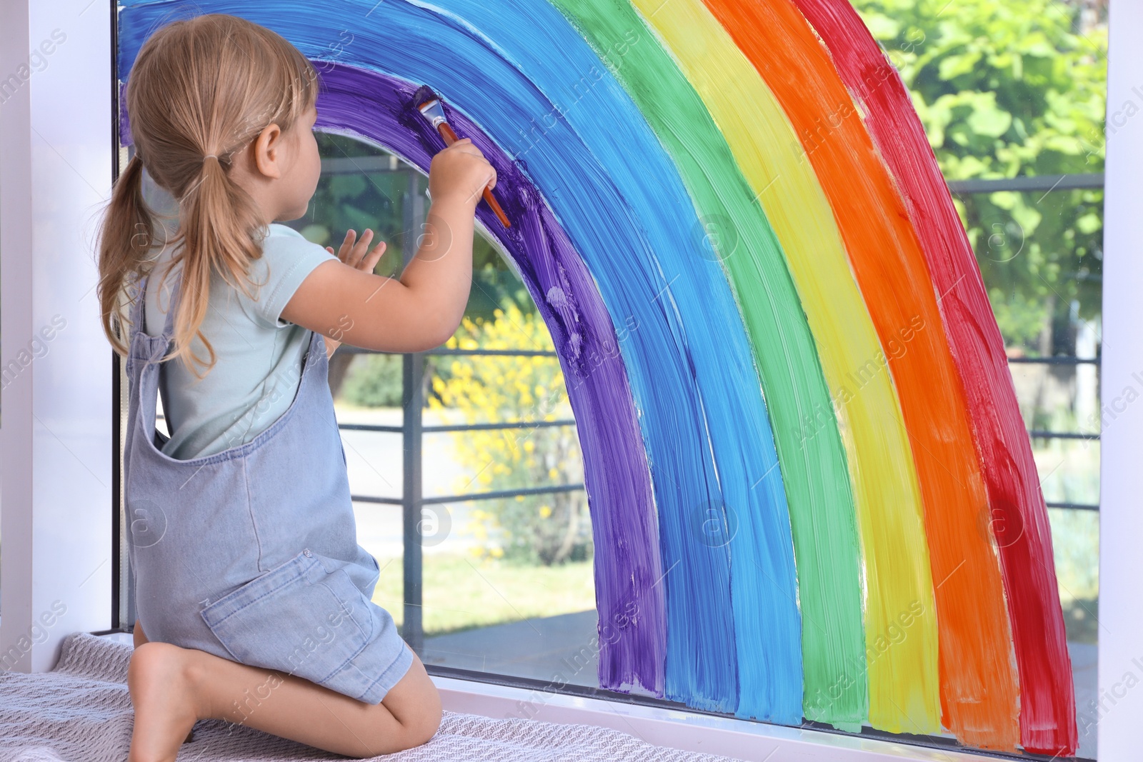 Photo of Little girl drawing rainbow on window indoors