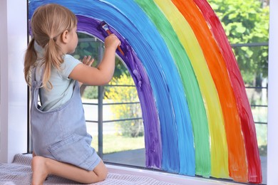 Little girl drawing rainbow on window indoors