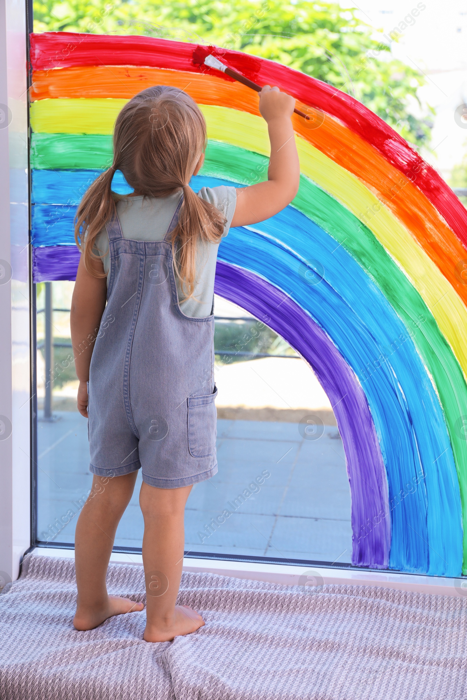 Photo of Little girl drawing rainbow on window indoors, back view