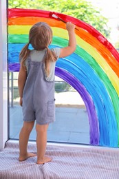 Little girl drawing rainbow on window indoors, back view