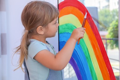 Little girl drawing rainbow on window indoors