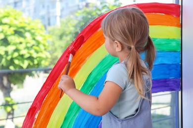 Little girl drawing rainbow on window indoors