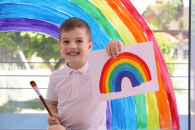Photo of Happy little boy with picture of rainbow and brush near window indoors
