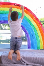 Little boy drawing rainbow on window indoors, back view