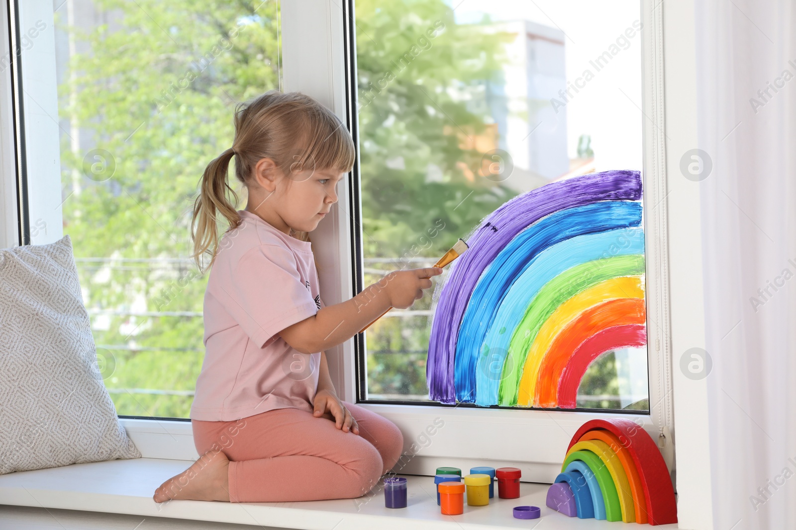 Photo of Little girl drawing rainbow on window indoors