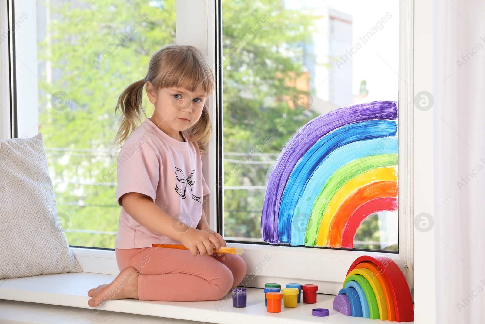 Photo of Little girl with brush near rainbow painting on window indoors