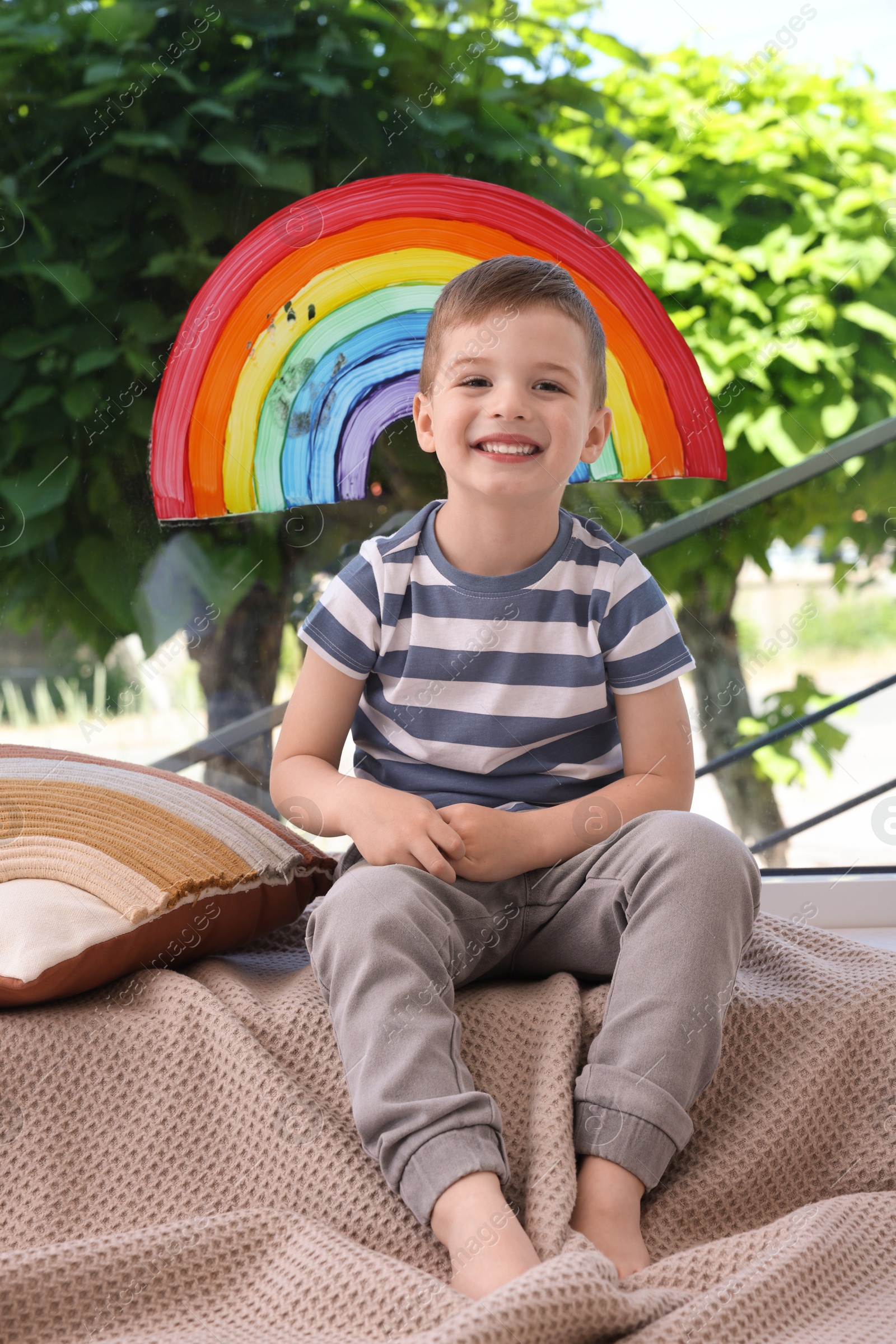 Photo of Happy little boy near rainbow painting on window indoors