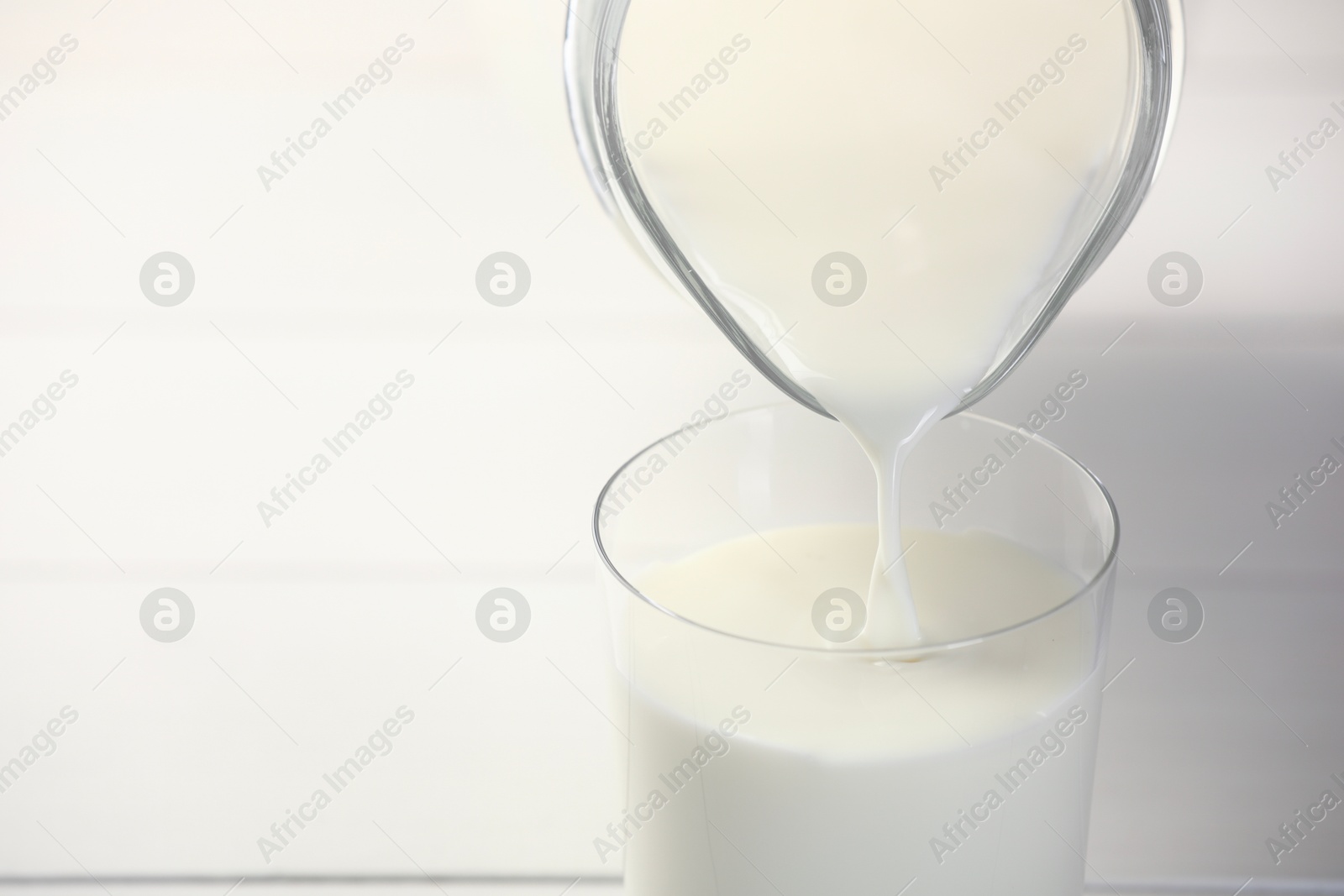 Photo of Pouring fresh milk from jug into glass at white table, closeup. Space for text