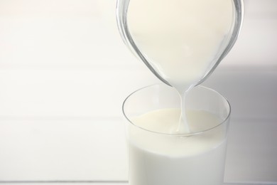 Pouring fresh milk from jug into glass at white table, closeup. Space for text