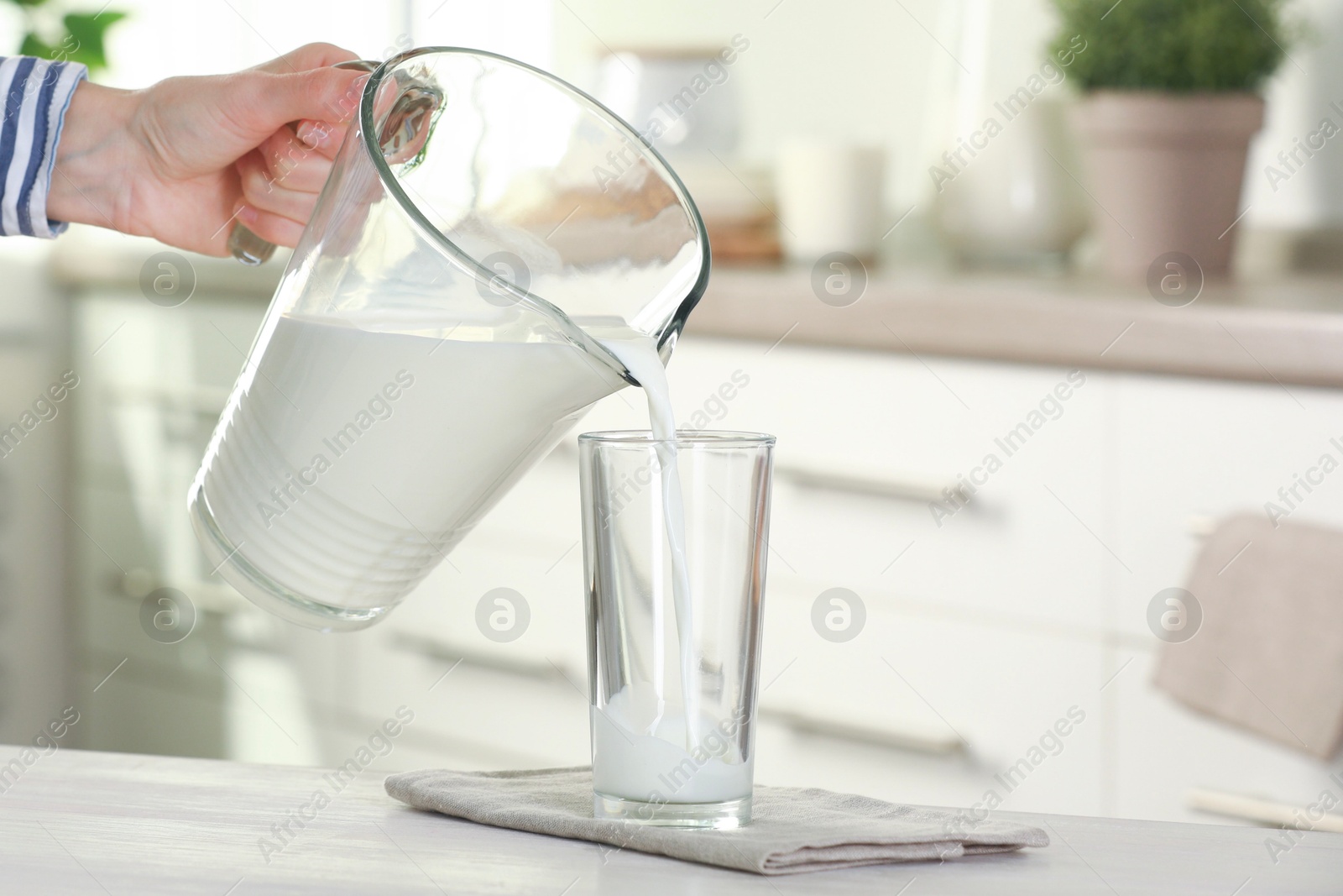 Photo of Woman pouring fresh milk from jug into glass at light wooden table in kitchen, closeup. Space for text