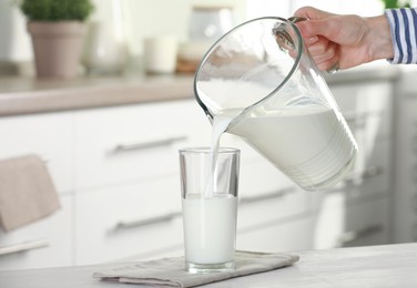 Photo of Woman pouring fresh milk from jug into glass at light wooden table in kitchen, closeup. Space for text