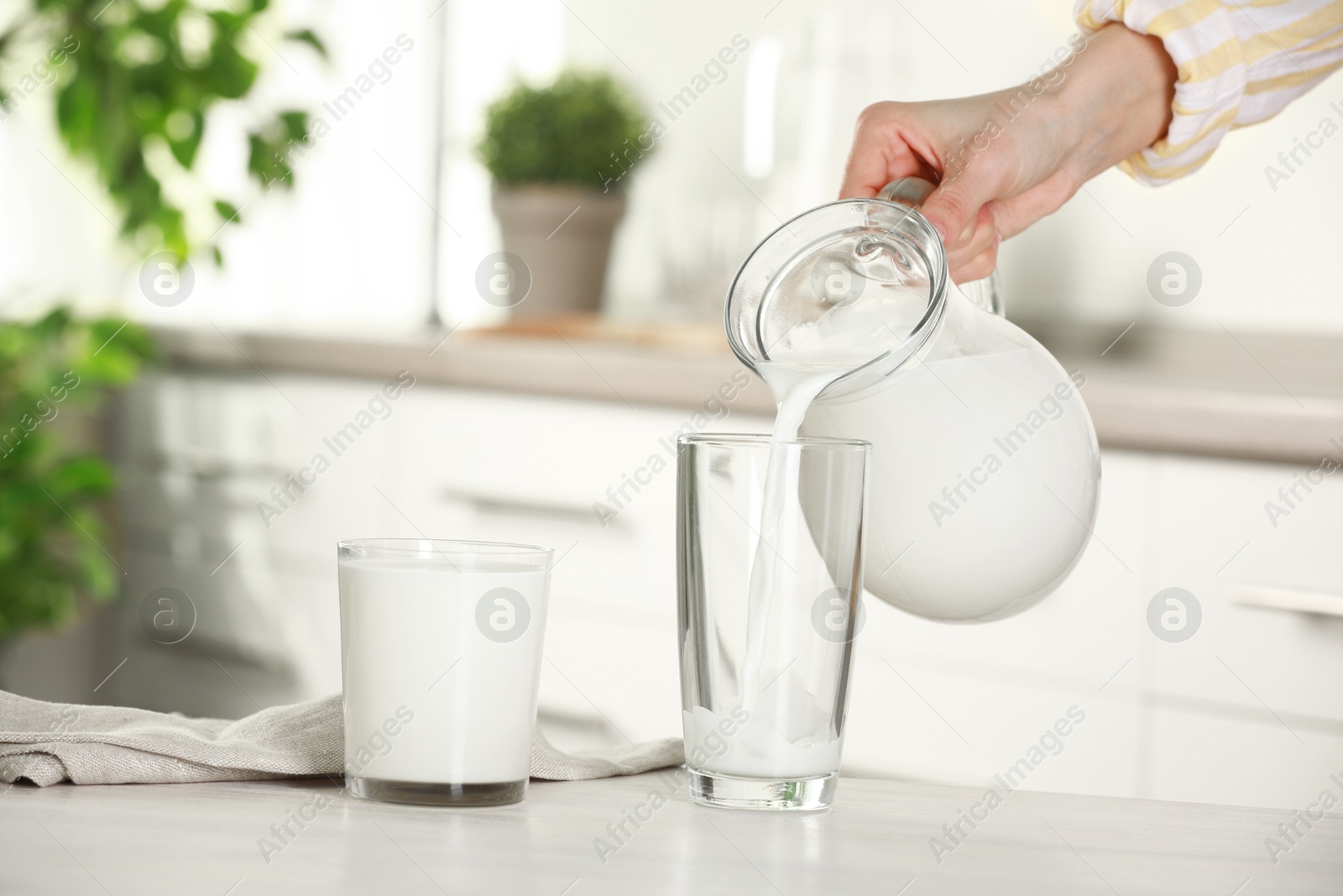 Photo of Woman pouring fresh milk from jug into glass at light wooden table in kitchen, closeup