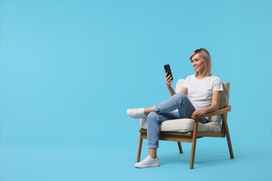 Happy woman with phone on armchair against light blue background, space for text
