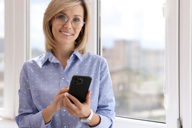 Happy woman using mobile phone near window indoors, space for text