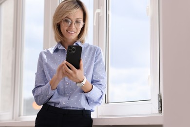 Happy woman using mobile phone near window indoors, space for text