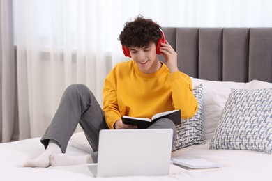 Teenager in headphones holding notebook and working with laptop on bed at home. Remote job