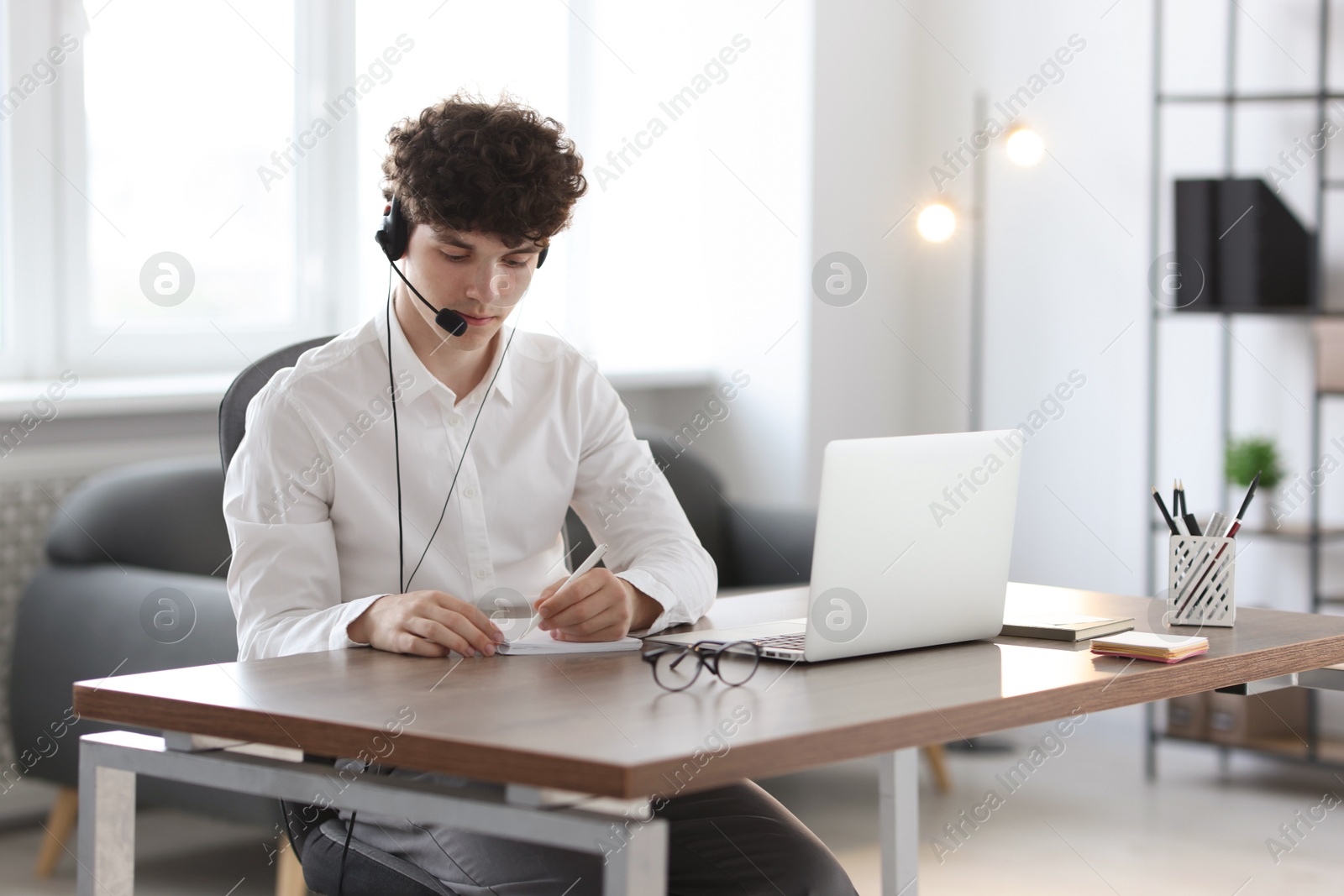 Photo of Teenager in headset taking notes while working with laptop at table indoors. Remote job