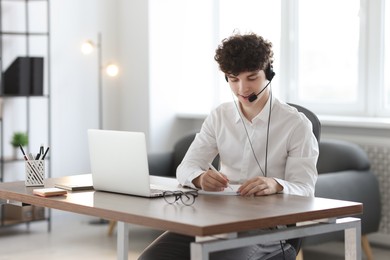 Photo of Teenager in headset taking notes while working with laptop at table indoors. Remote job