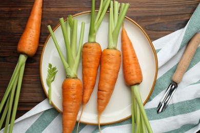 Fresh carrots and vegetable peeler on wooden table, flat lay