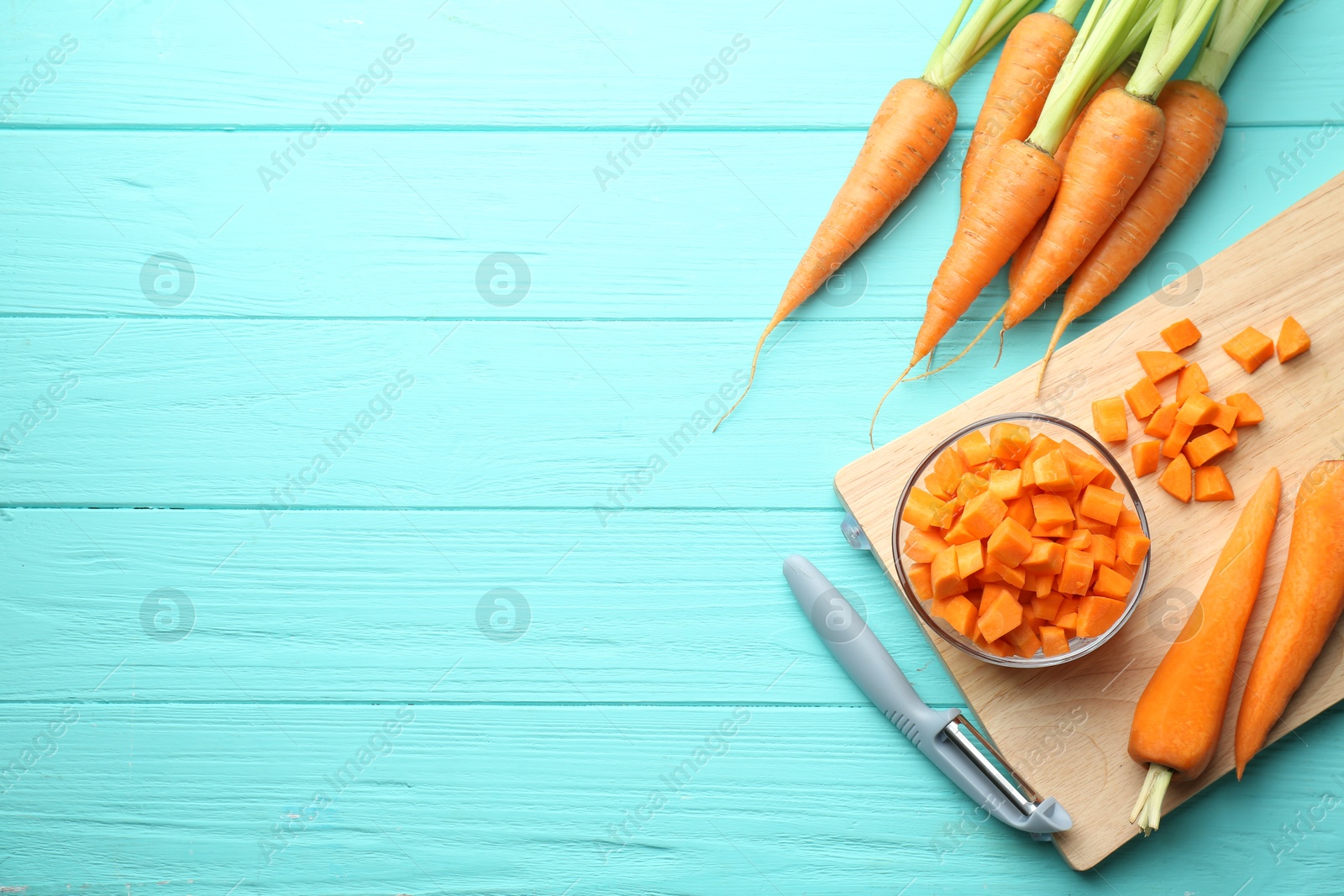 Photo of Chopped carrot in bowl, fresh vegetables and peeler on light blue wooden table, flat lay. Space for text