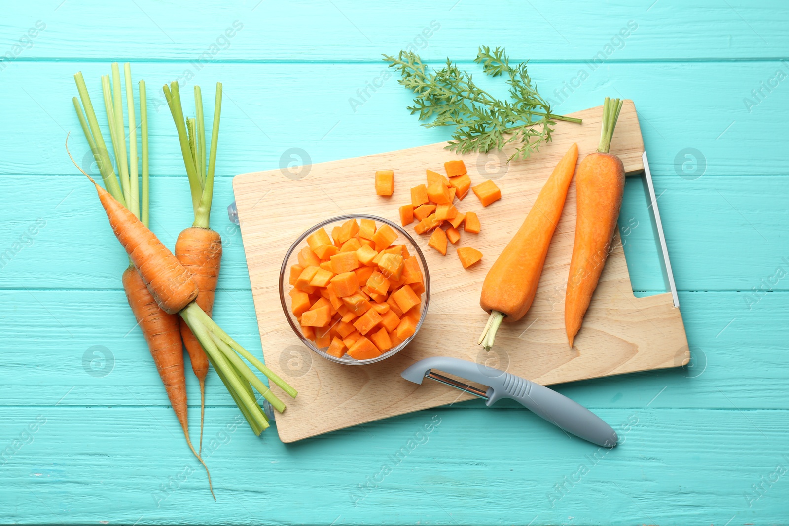 Photo of Chopped carrot in bowl, fresh vegetables and peeler on light blue wooden table, flat lay