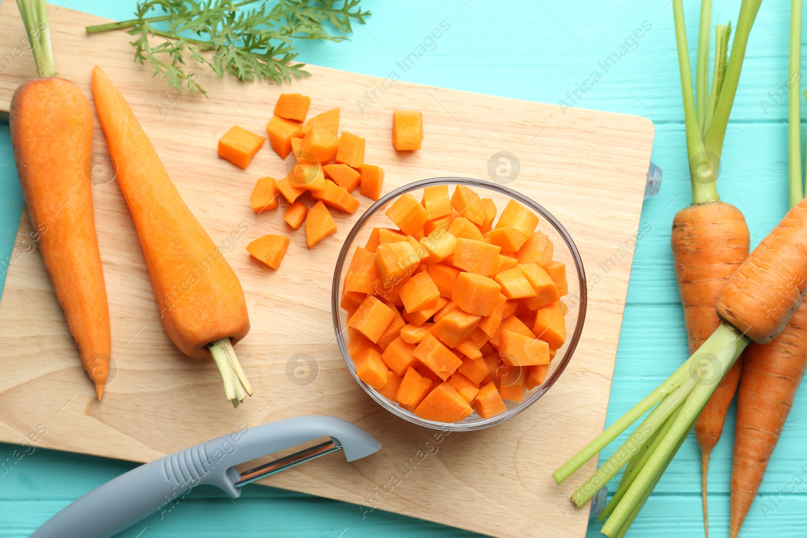 Photo of Chopped carrot in bowl, fresh vegetables and peeler on light blue wooden table, flat lay