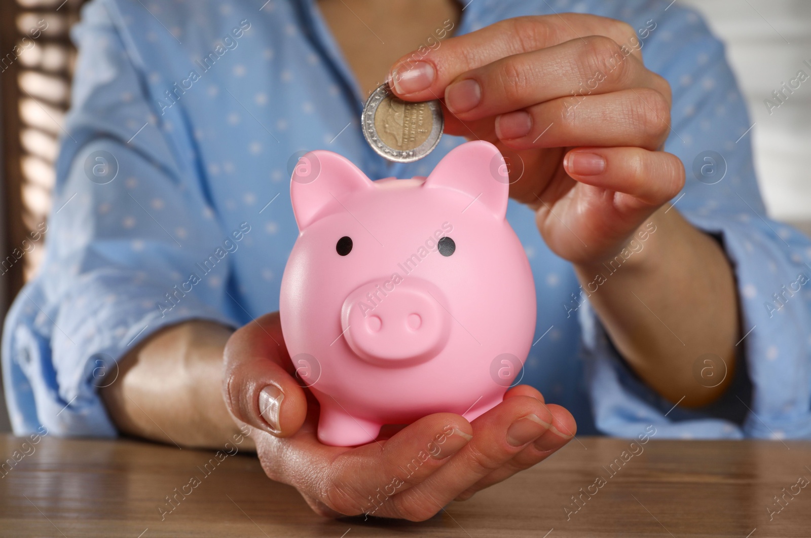 Photo of Woman putting coin into pink piggy bank on wooden table, closeup