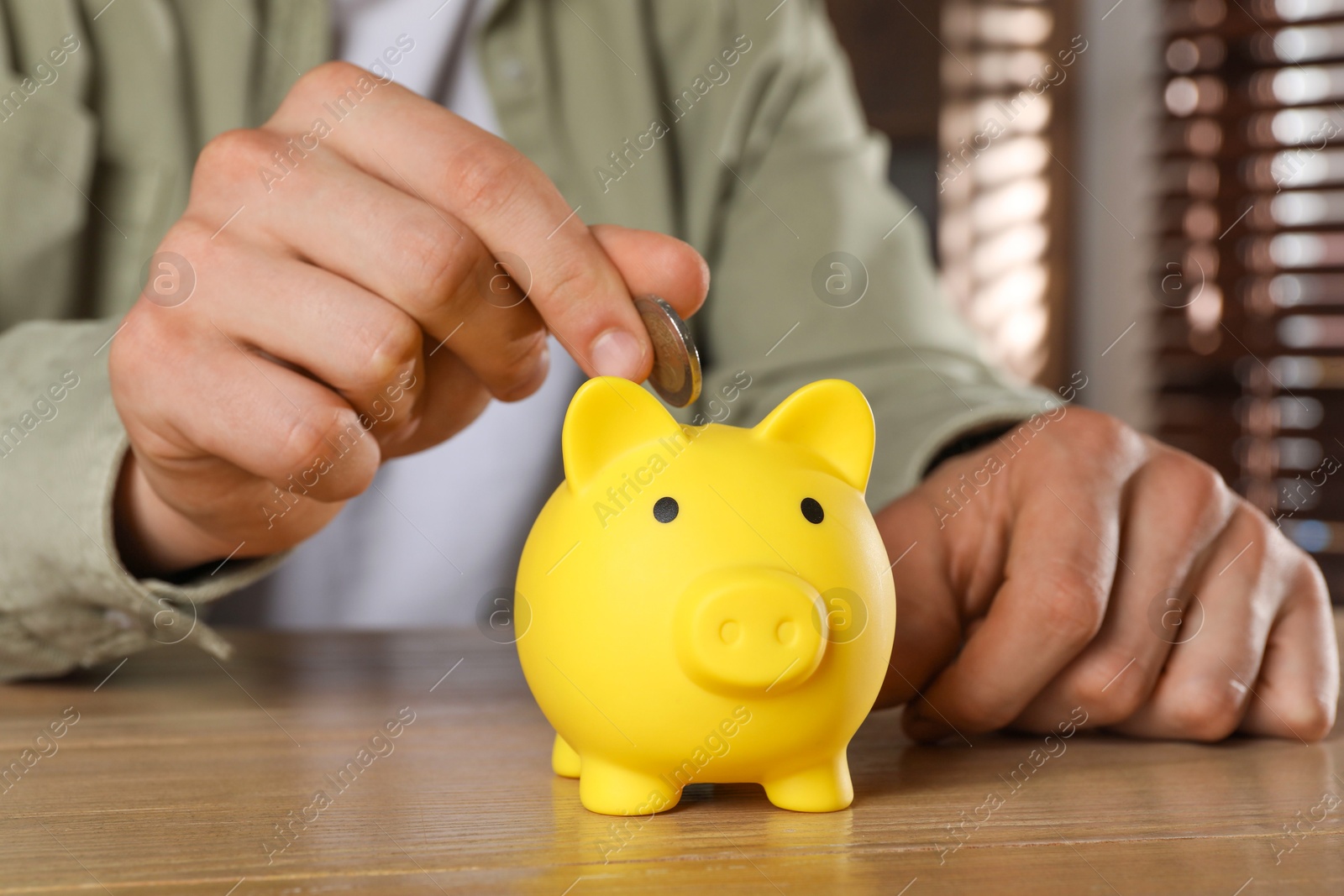 Photo of Man putting coin into yellow piggy bank on wooden table, closeup