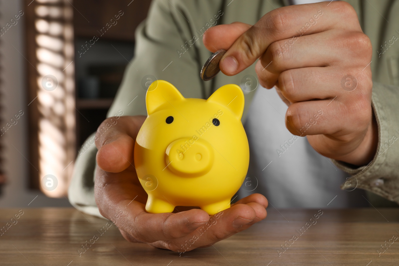 Photo of Man putting coin into yellow piggy bank on wooden table, closeup