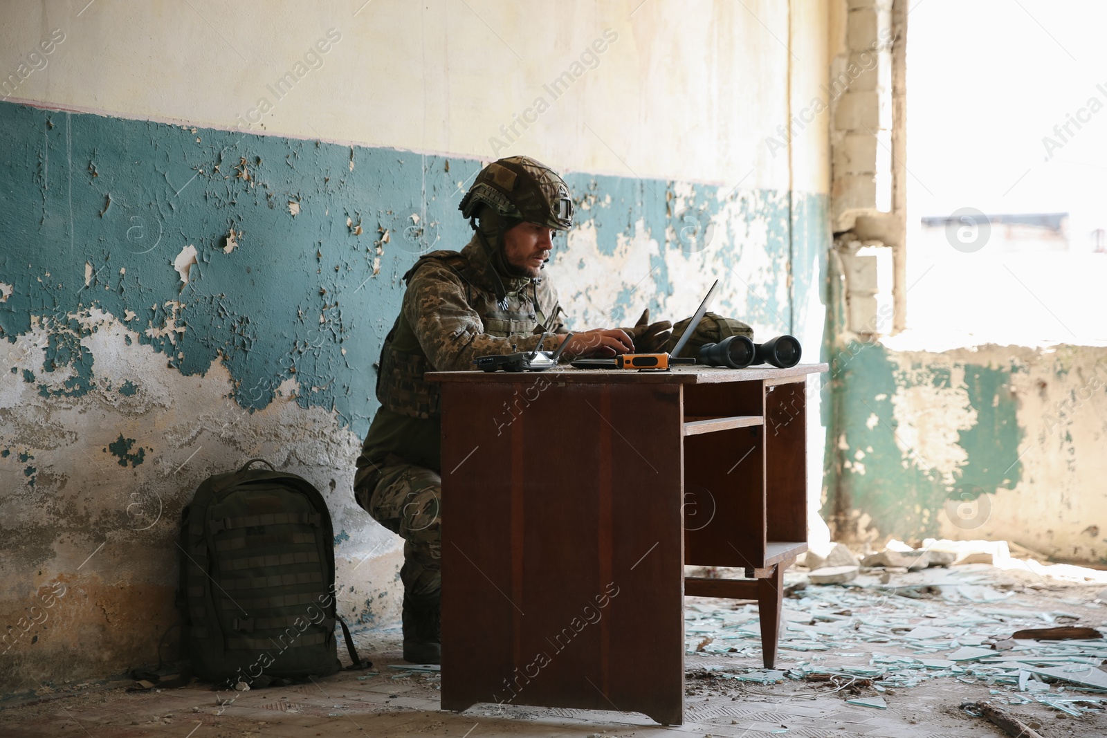 Photo of Military mission. Soldier in uniform using laptop at table inside abandoned building