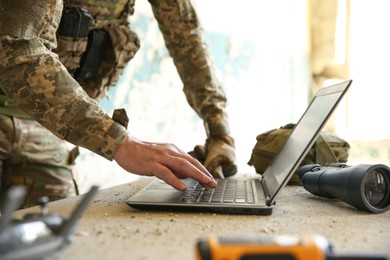 Military mission. Soldier in uniform using laptop at table inside abandoned building, closeup