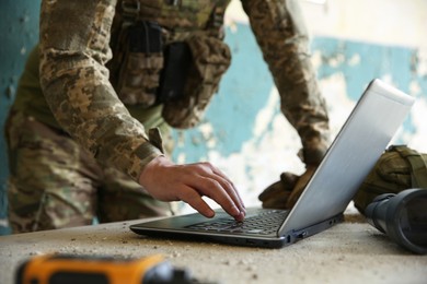 Military mission. Soldier in uniform using laptop at table inside abandoned building, closeup