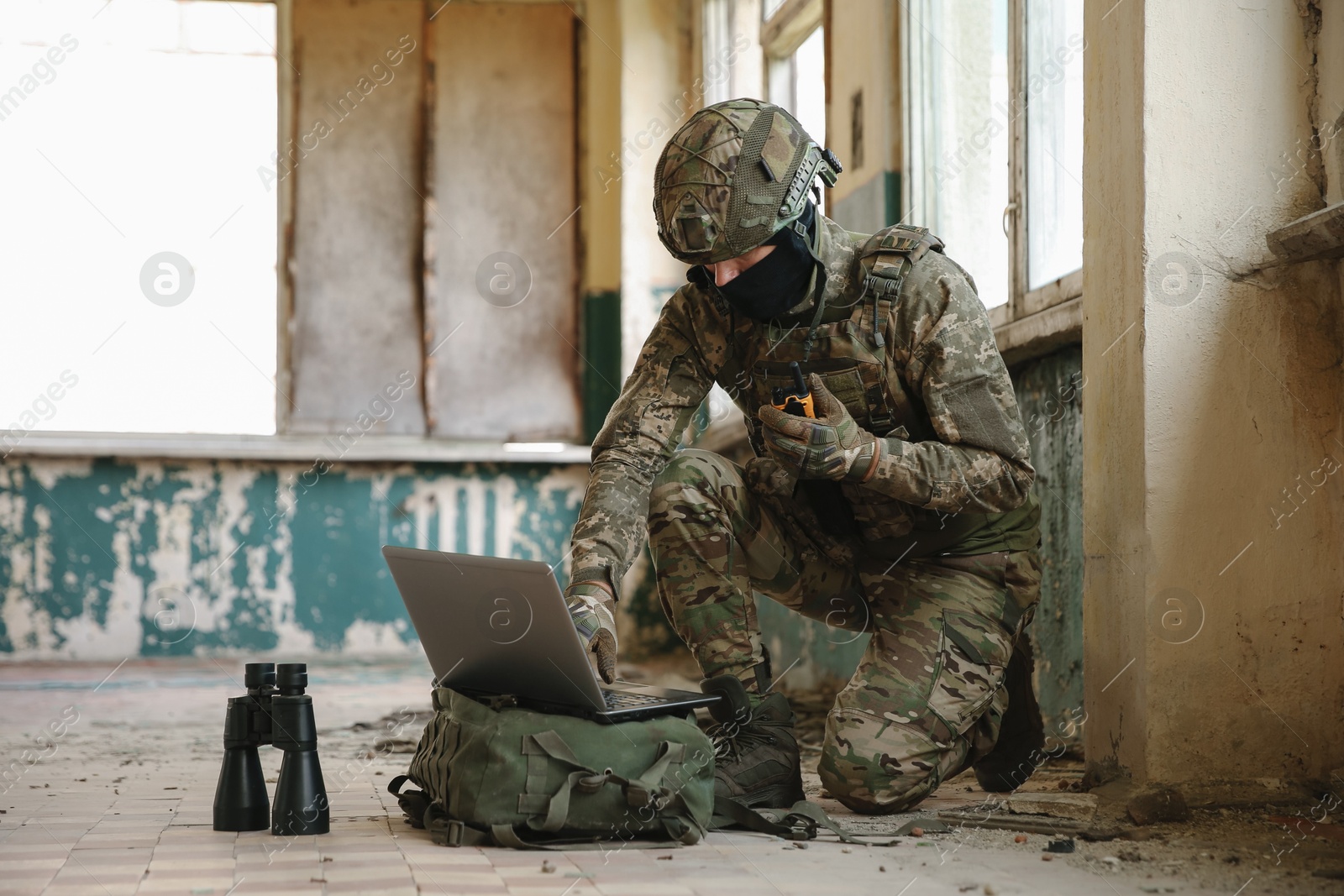 Photo of Military mission. Soldier in uniform using laptop and binoculars inside abandoned building