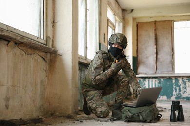 Military mission. Soldier in uniform using laptop and binoculars inside abandoned building