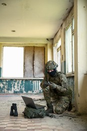 Military mission. Soldier in uniform using laptop and binoculars inside abandoned building