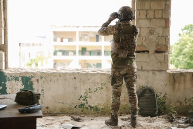 Military mission. Soldier in uniform with binoculars inside abandoned building, back view. Space for text