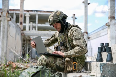 Photo of Military mission. Soldier in uniform using laptop near abandoned building outside